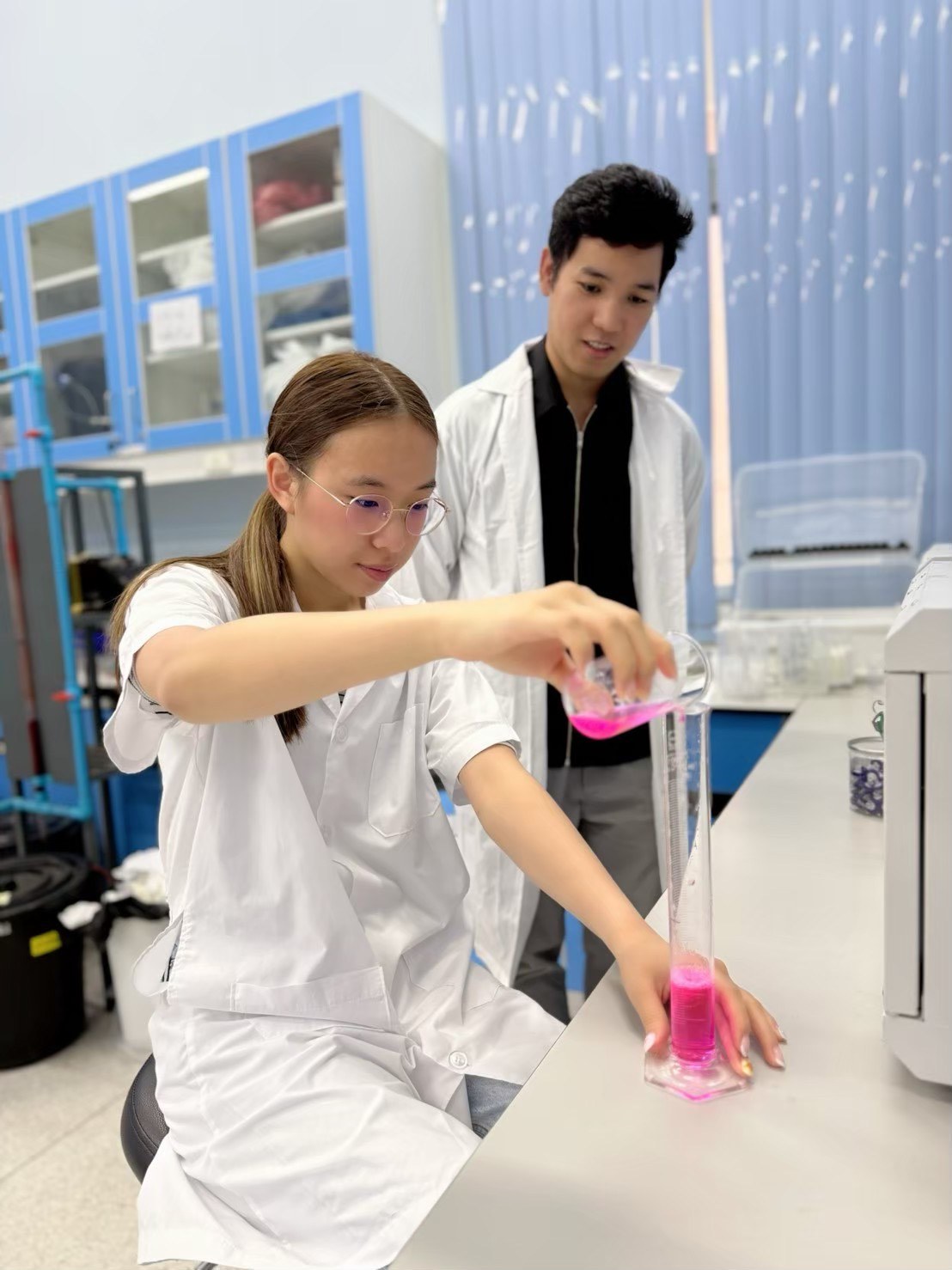 Winnie pouring a pink sample into a measuring cylinder while a researcher looks on